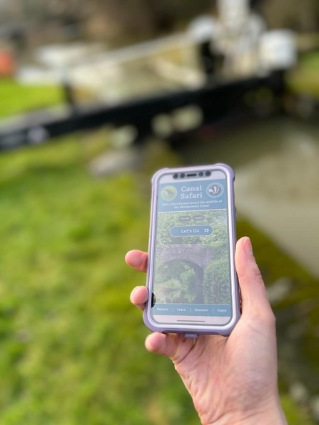 A hand holding a smartphone with Canal Safari app open and a canal lock in the background