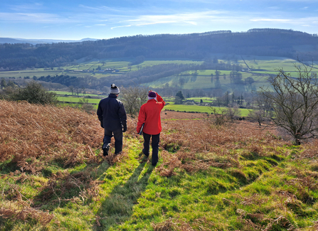 Two people in winter clothes, standing with their backs to camera, stand looking over some bracken habitat. There is a tree covered hill in the background