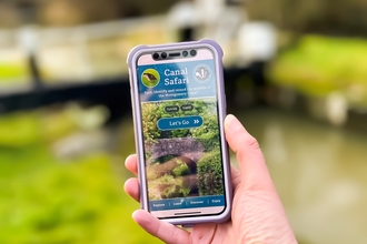 Hand holding a smartphone in front of a canal, with a canal lock in the background