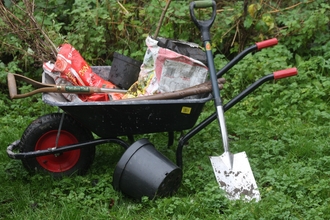 A black wheelbarrow with red handles, with bags of saplings, a spade and plant pots in it. There is a spade leaning against the wheelbarrow and another plant pot on the ground in front.