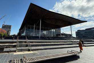 Exterior view of the Senedd in Cardiff Bay, showing its distinctive wide, overhanging roof and glass façade, with steps and a few people in the foreground under a bright blue sky with scattered clouds.
