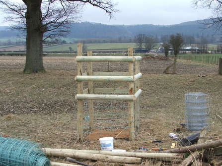 A photo showing a single tree planted inside a small fenced exclosure (post and rail). At the front of the image are unused fencing materials across the ground. Behind the fenced tree, to the left middle distance is a mature tree and beyond are sheep grazed fields and distant hills covered in trees. It is winter.