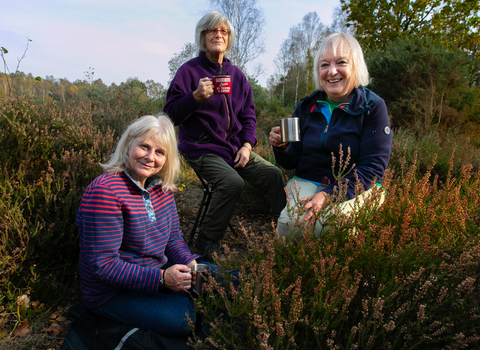 Three older ladies sitting outside drinking tea, all looking at the camera and smiling