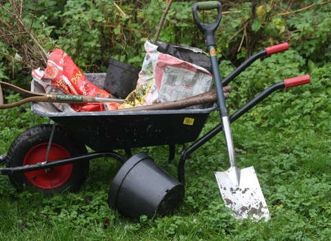 A black wheelbarrow with red handles, with bags of saplings, a spade and plant pots in it. There is a spade leaning against the wheelbarrow and another plant pot on the ground in front.