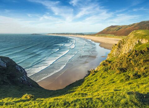 A wide coastal landscape viewed from a grassy cliff, overlooking a long sandy beach with gentle waves rolling in from the blue ocean. Rolling green hills and rocky cliffs frame the shoreline under a partly cloudy sky.